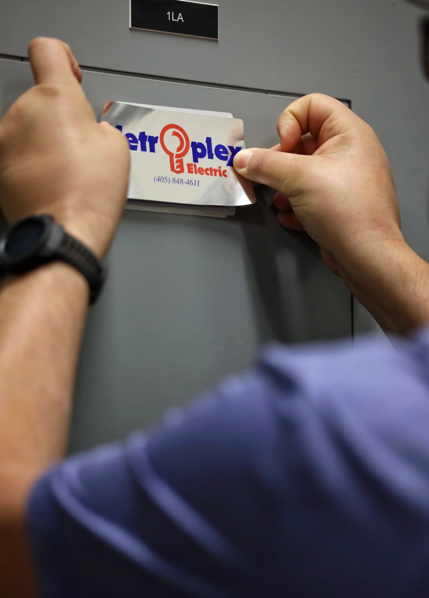 An electrician's hands applying a Metroplex label to a finished grey electrical panel